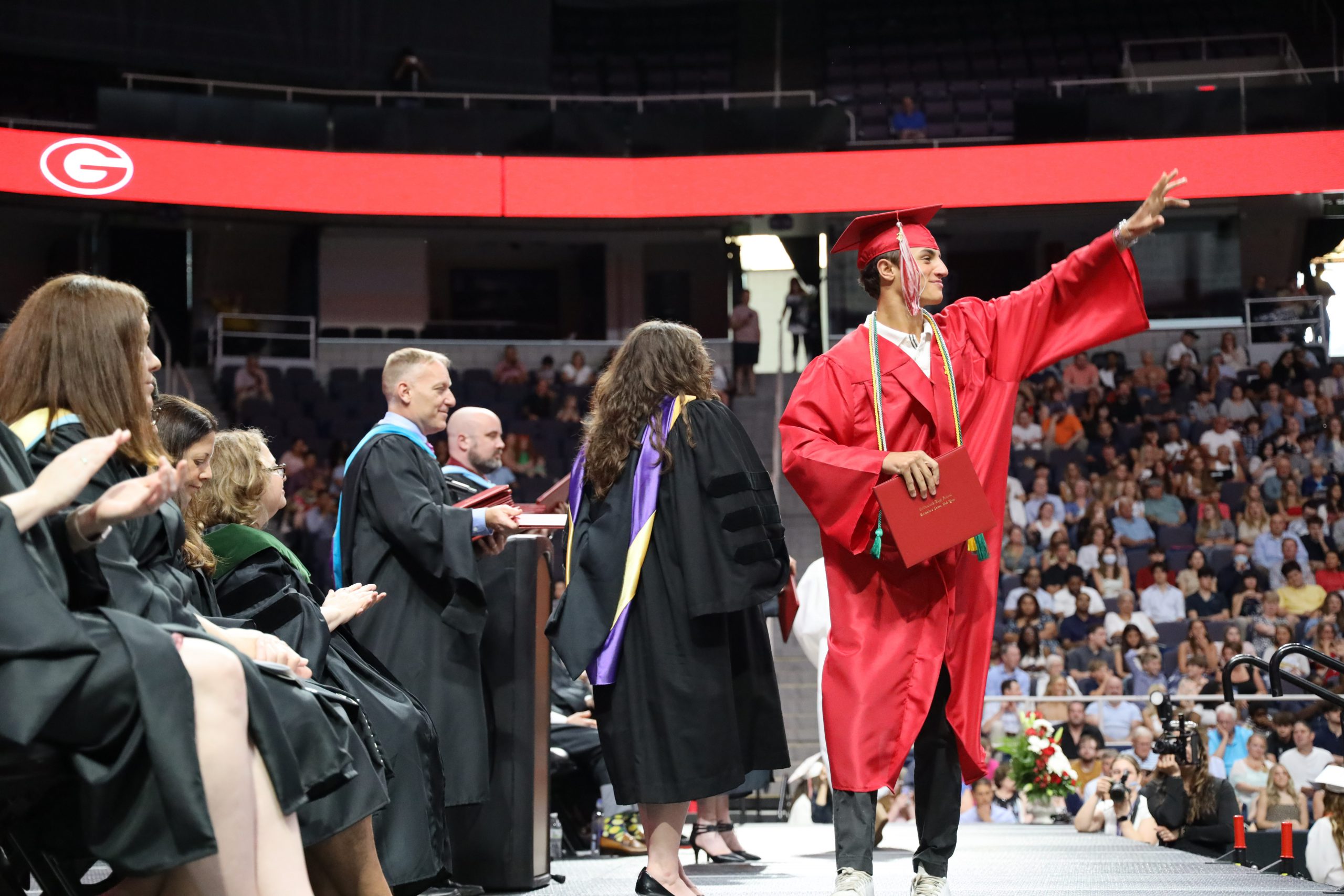 A students wearing a red cap and gown waves at the crowd after just receiving a diploma