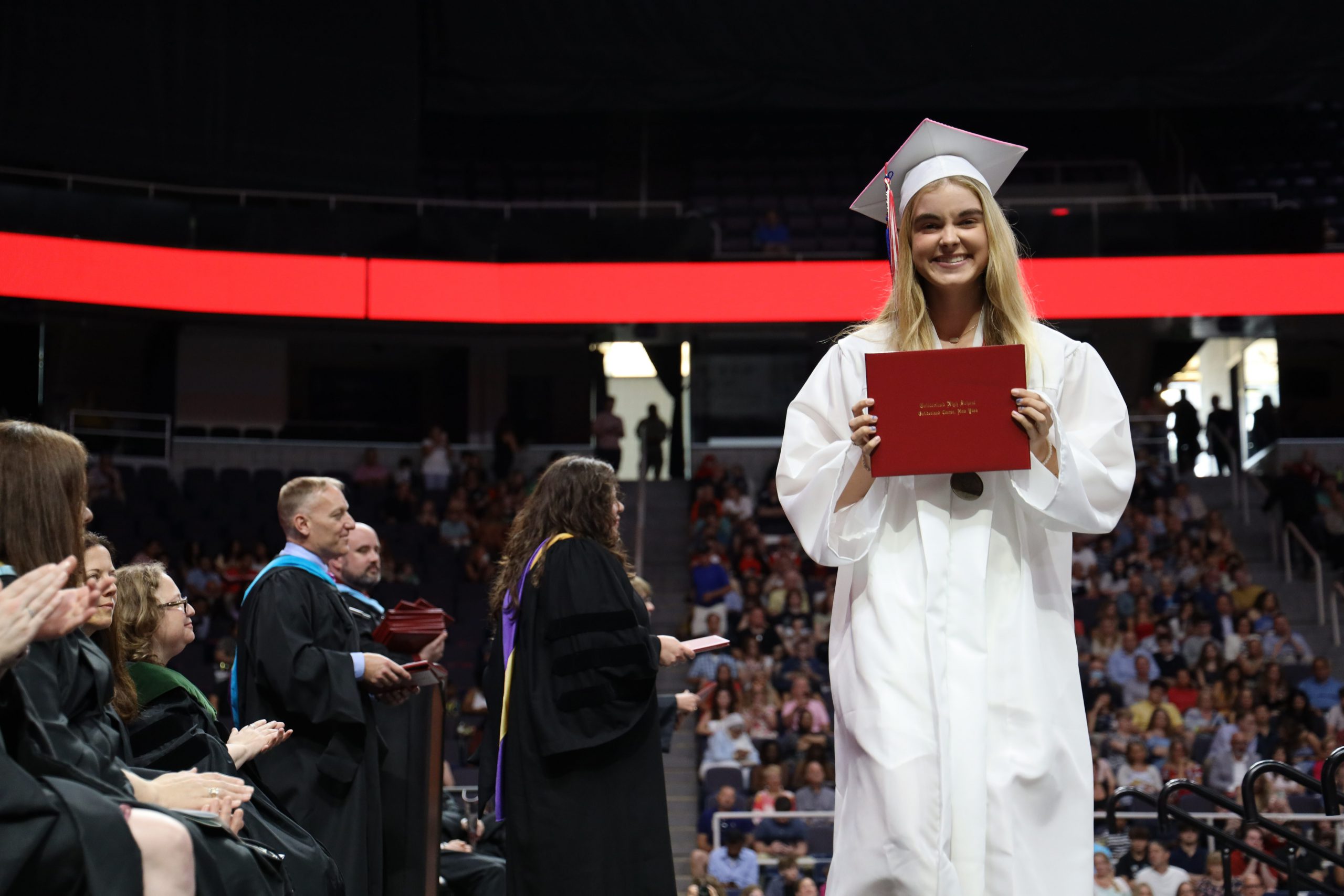 A student wearing a white cap and gown holds up a diploma and is smiling