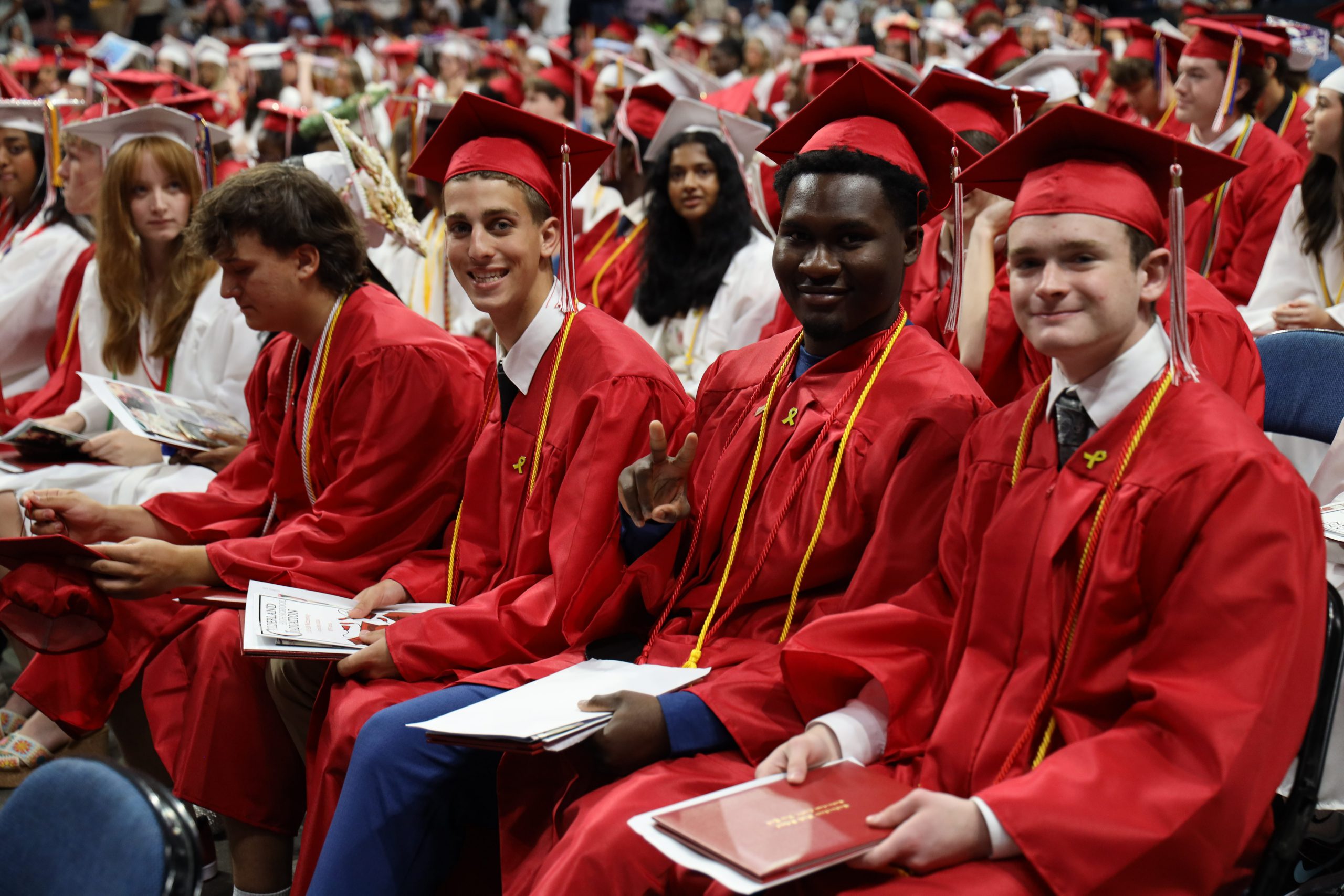 Students wearing caps and gowns, some are red and some are white, are seated in rows. A few are looking at the camera and smiling. One is giving a peace sign
