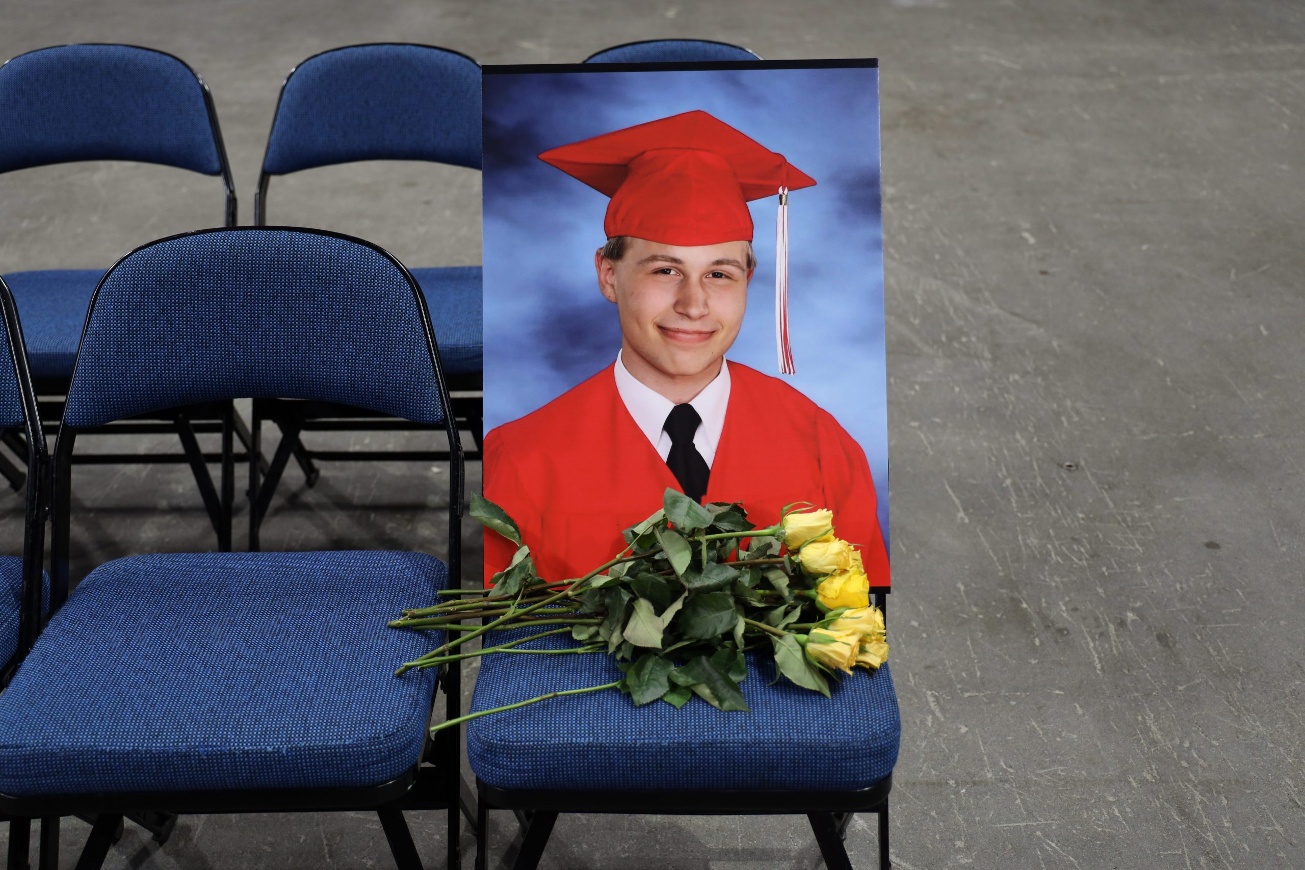 A blue chair at the end of a row of chairs with a picture of a graduate in red cap and gown. A bouquet of yellow roses sits in front of the picture