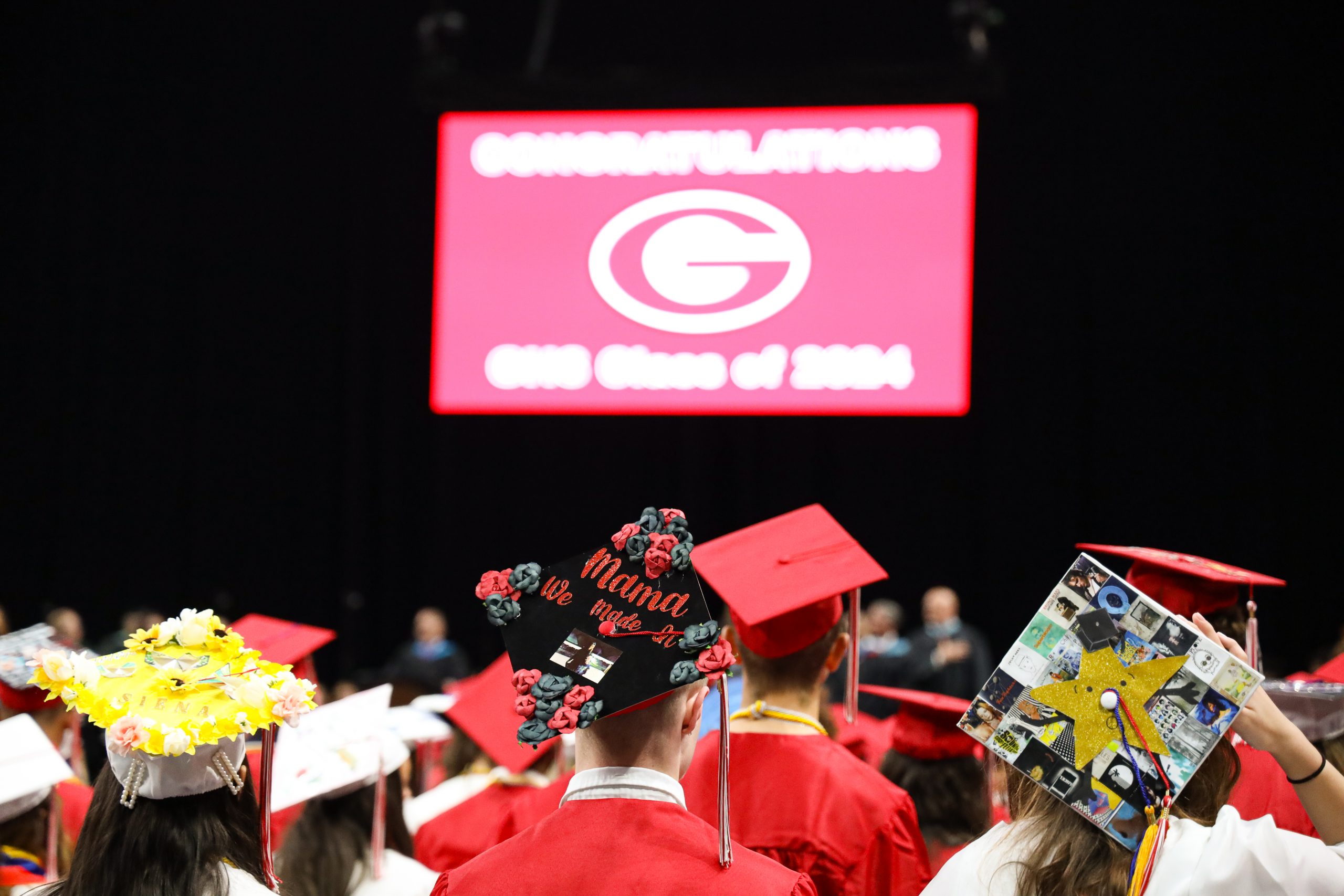 In the background is a screen with a large red G. In the foreground are three students with decorated caps