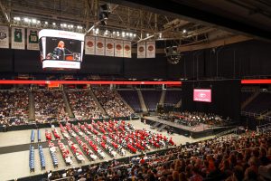 A wide shot of graduation at MVP Arena. There are rows of chairs on the floor of the arena. GHS students are wearing red robes and white gowns. The stage is to the right