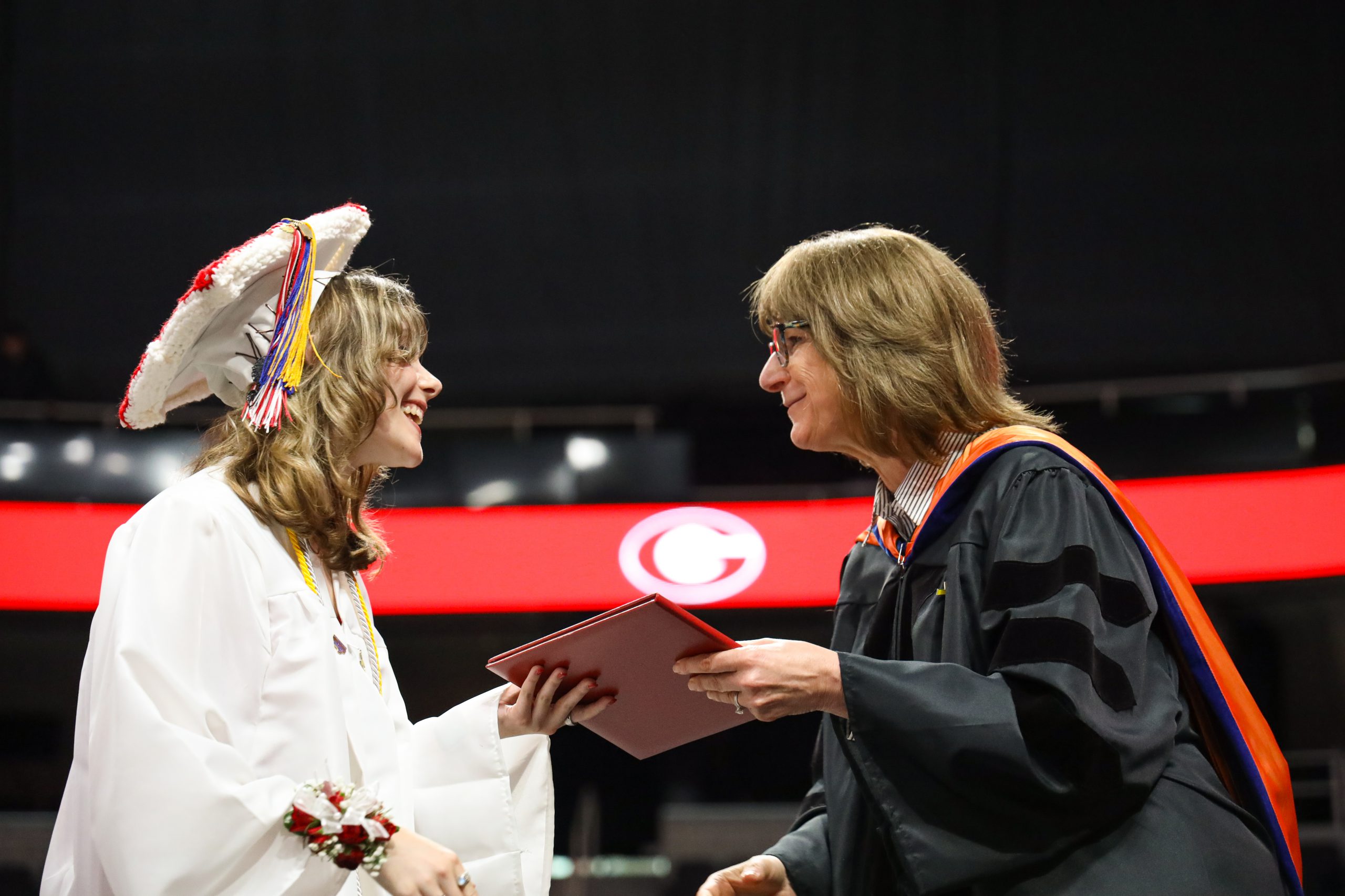 A students wearing a white cap and gown accepts her diploma from the superintendent who is wearing a black gown with an orange hood