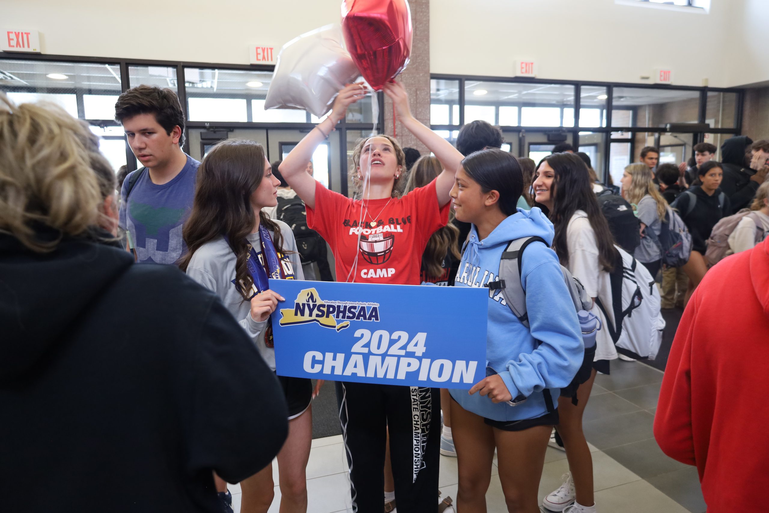 Two students are holding a sign that reads NYSPHSAA 2024 Champion. In between them is a student holding balloons
