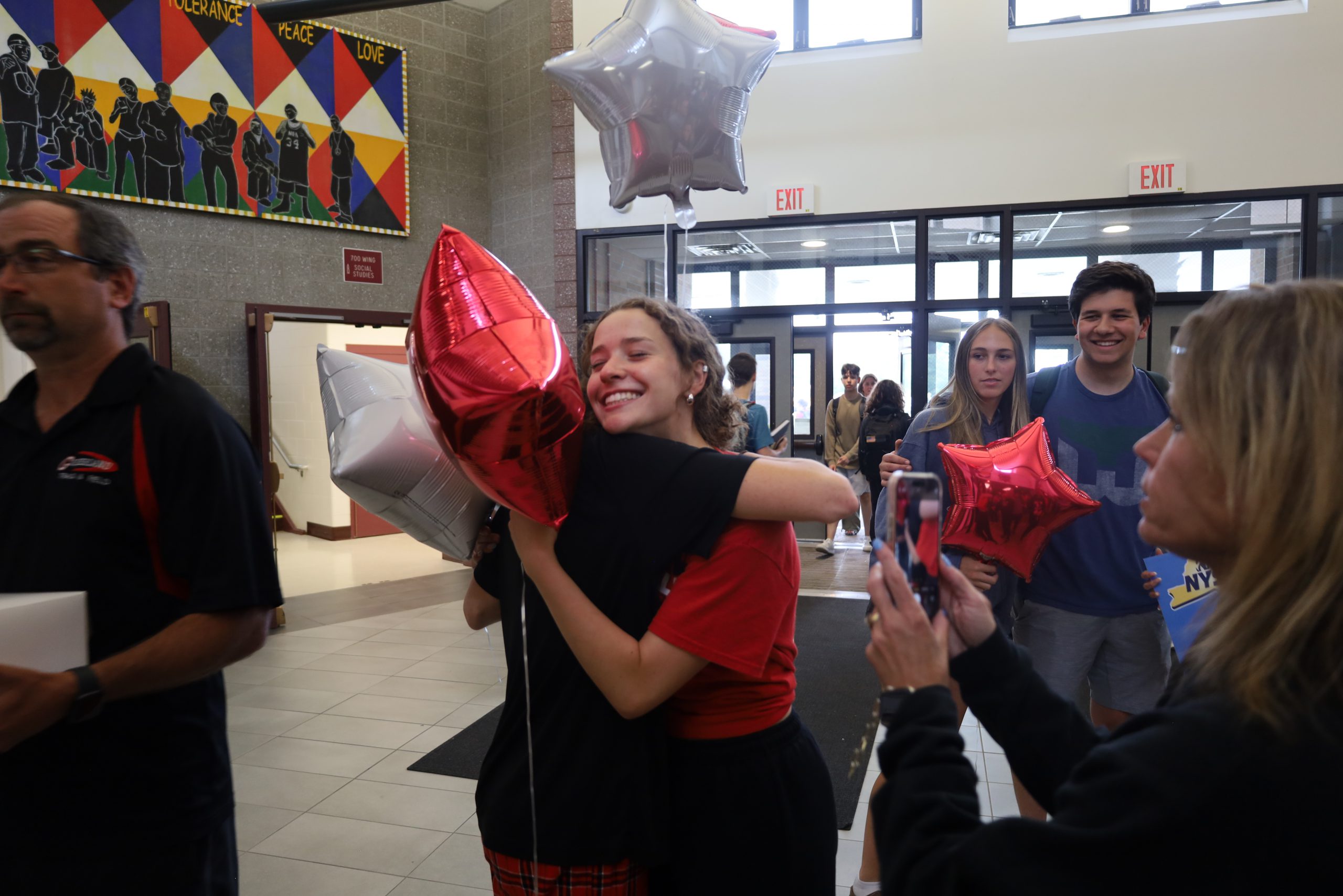 A student hugs another students. One is holding a red balloon in the shape of a star