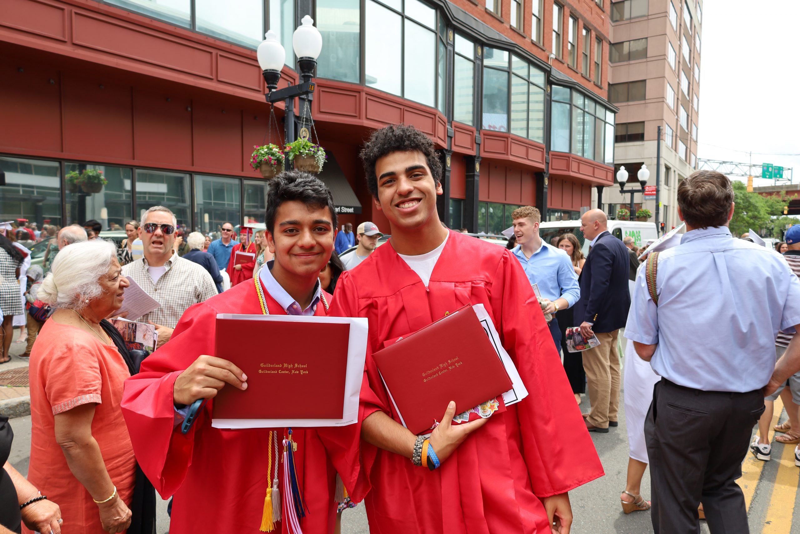Two students wearing red gowns hold up their diplomas. They are standing side by side outside, people are waling behind them