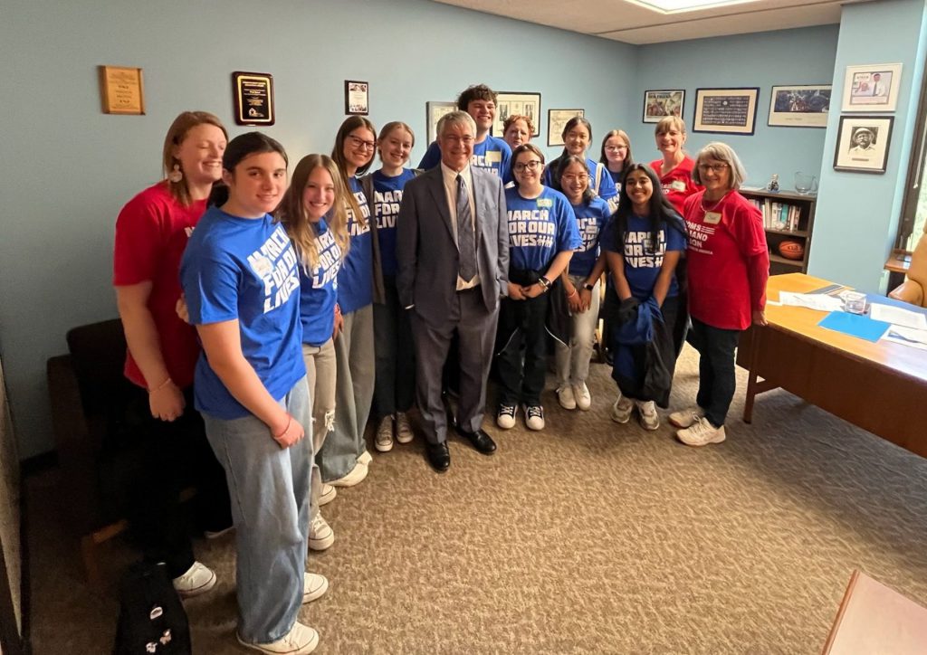 Students wearing blue shirts standing for a group photo with a legislator, who's dressed in a gray suit and standing in the middle. Two women wearing red shirts are standing on the right side of the group, while another woman is on the left side. Everyone is smiling at the camera.