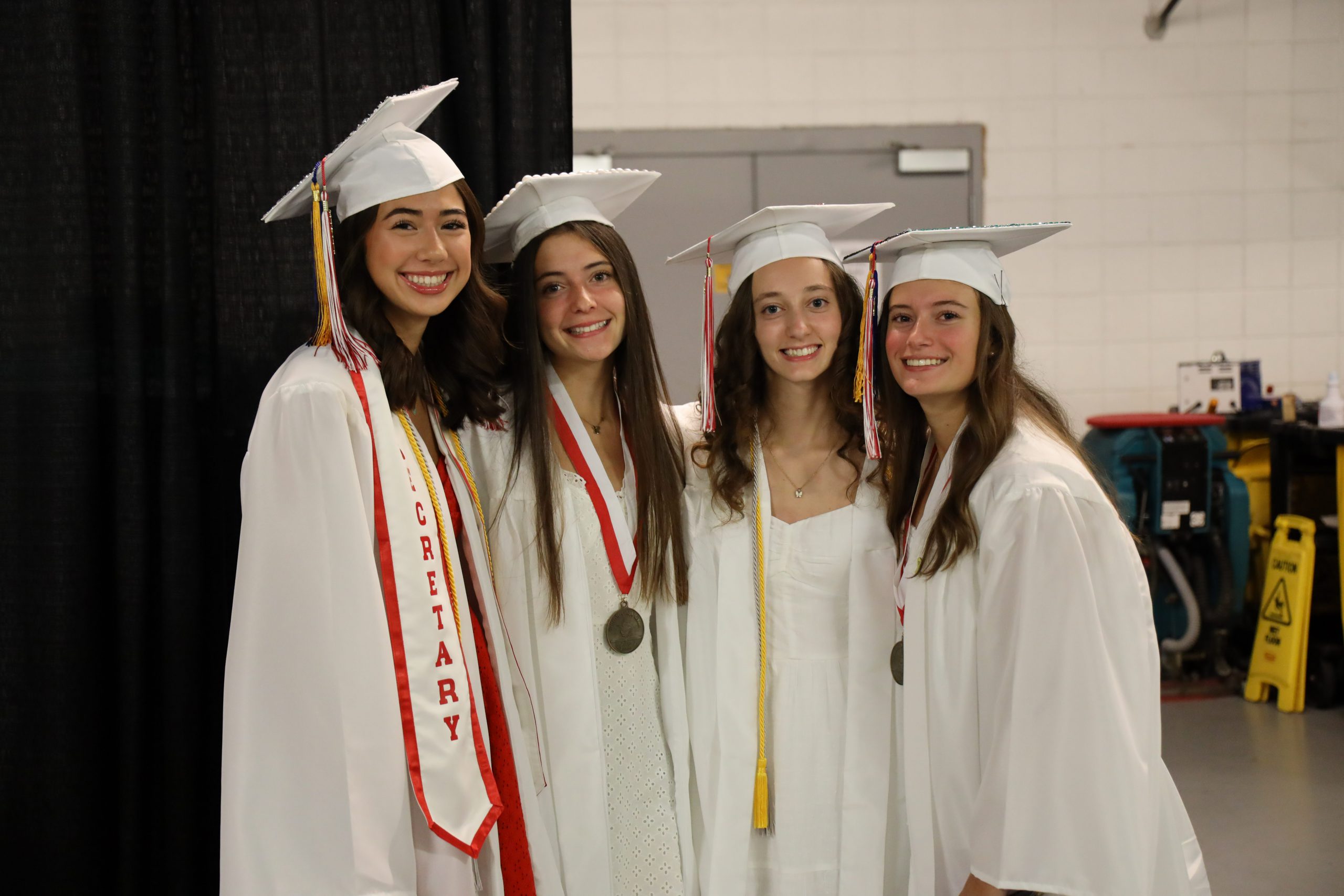 Four female graduates wearing white caps and gowns are smiling for a group photo together.