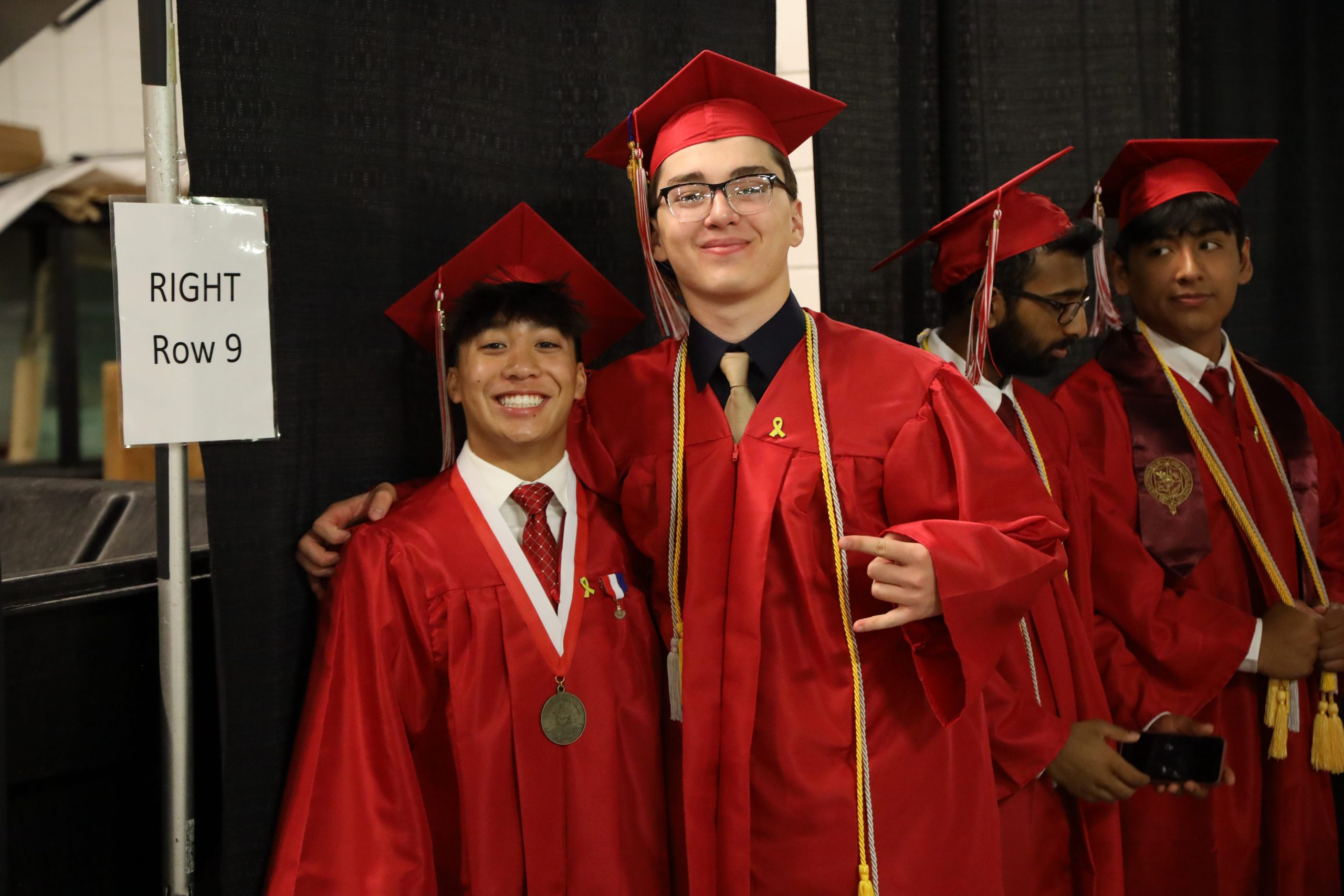 Two graduates wearing red caps and gowns smiling for a photo together. The boy on the left is wearing a medal around his neck, while the boy on the right is wearing a yellow cord around his.