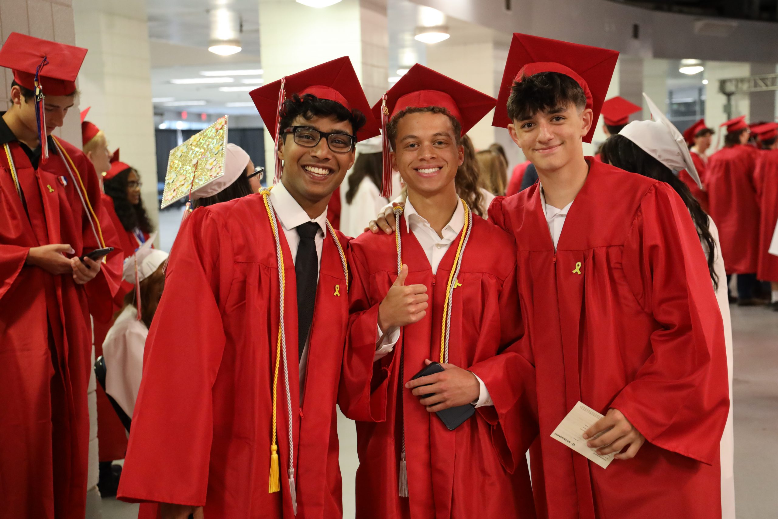 Three graduates wearing red caps and gowns are smiling together for a group photo. The boy in the middle is giving a thumbs up.