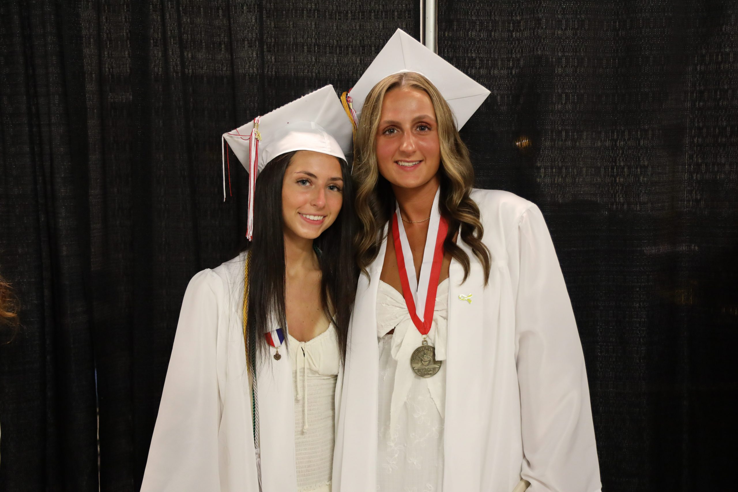 Two graduating students smiling together for a photo. Both girls are wearing white caps and gowns. The graduate on the right is also wearing a medal around her neck.