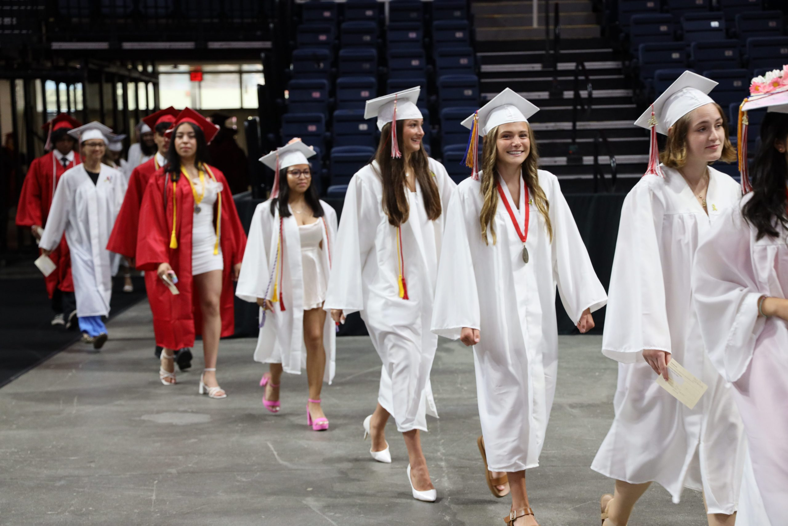 Group of graduates walking into the arena. Most of the graduates are wearing white caps and gowns, while a few students in the back of the photo are wearing red caps and gowns.