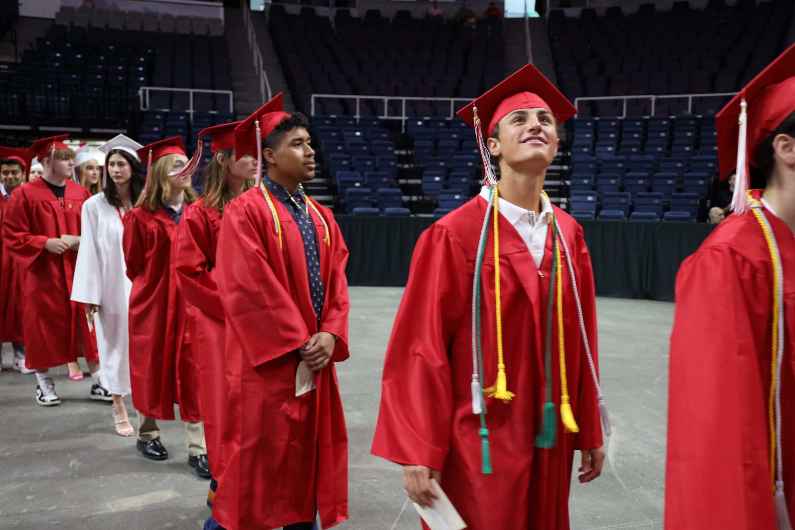 Graduates walking in to the arena. The first graduate is looking up as he walks in. Most of the graduates are wearing red caps and gowns, while one student in the very back of the photo is wearing a white cap and gown.