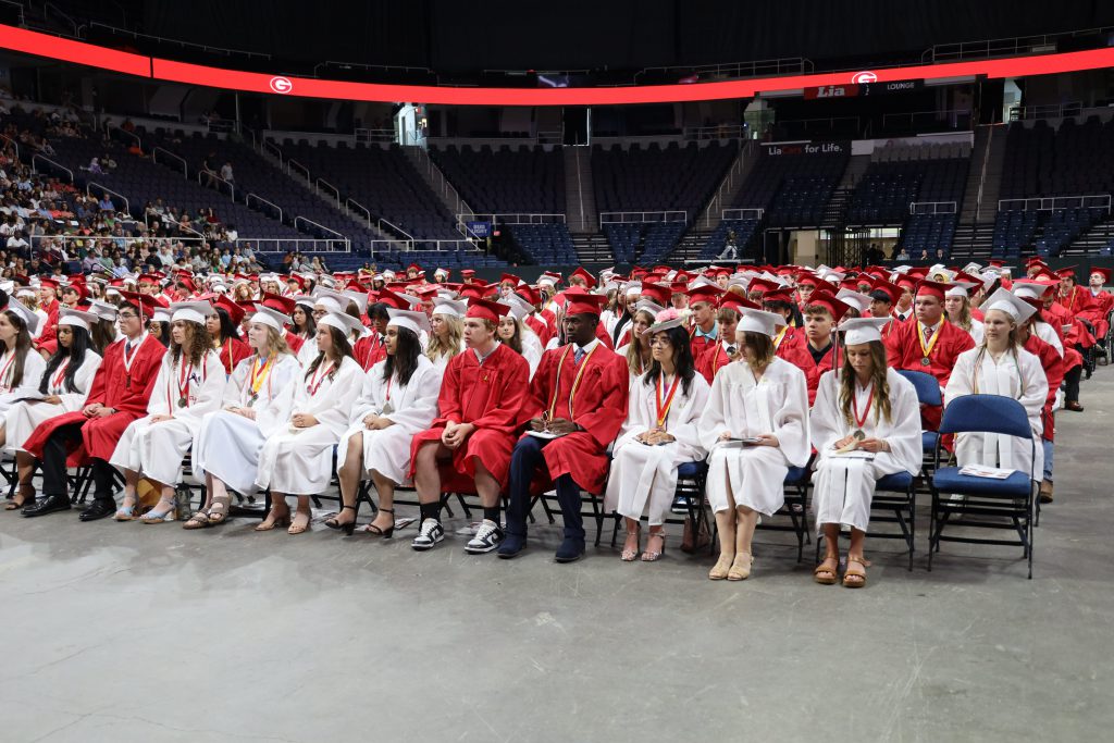 Audience of graduating students sitting in their seats in an arena. All are dressed in red or white caps and gowns.