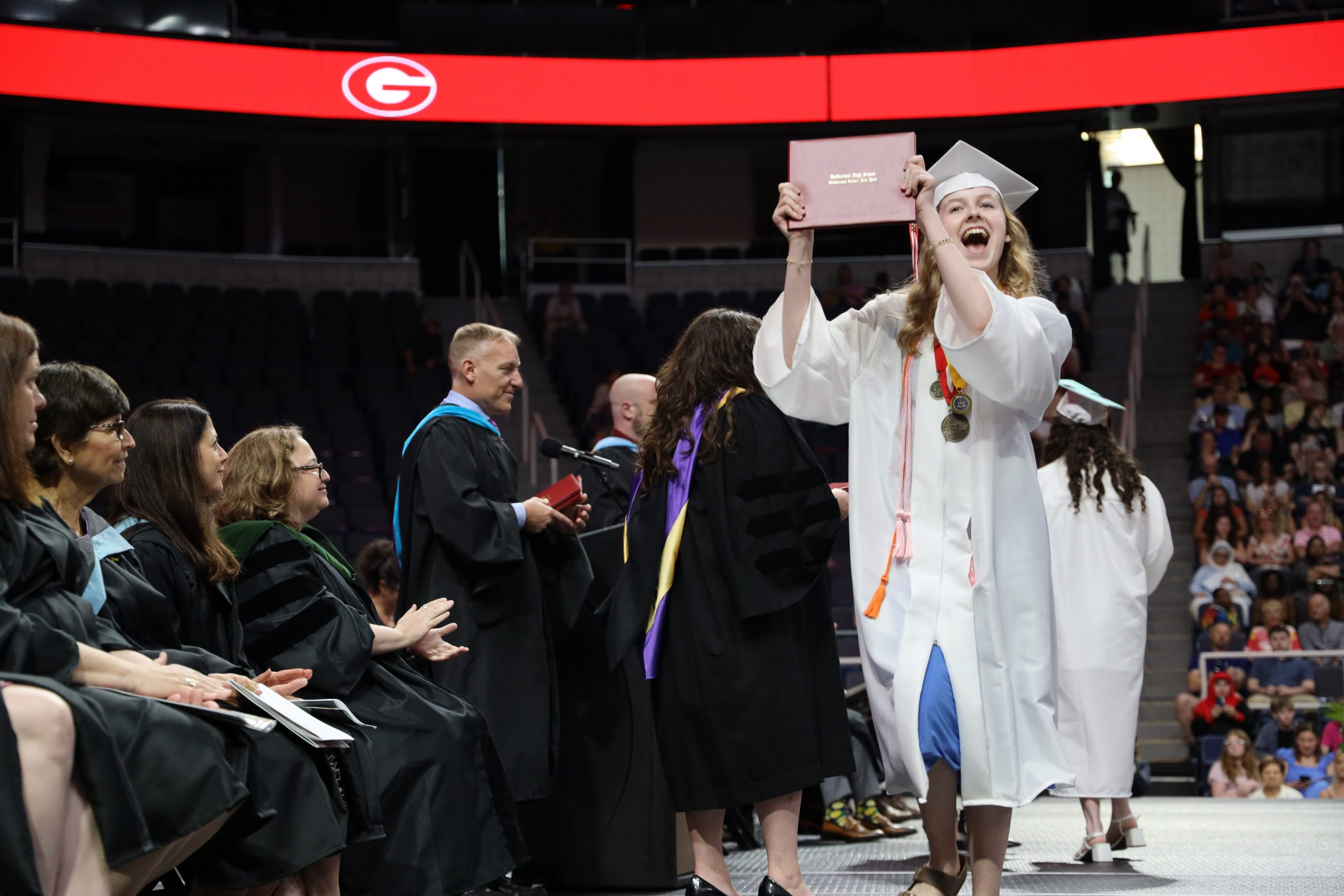 Graduate in a white cap and gown holding up her diploma on stage and smiling.