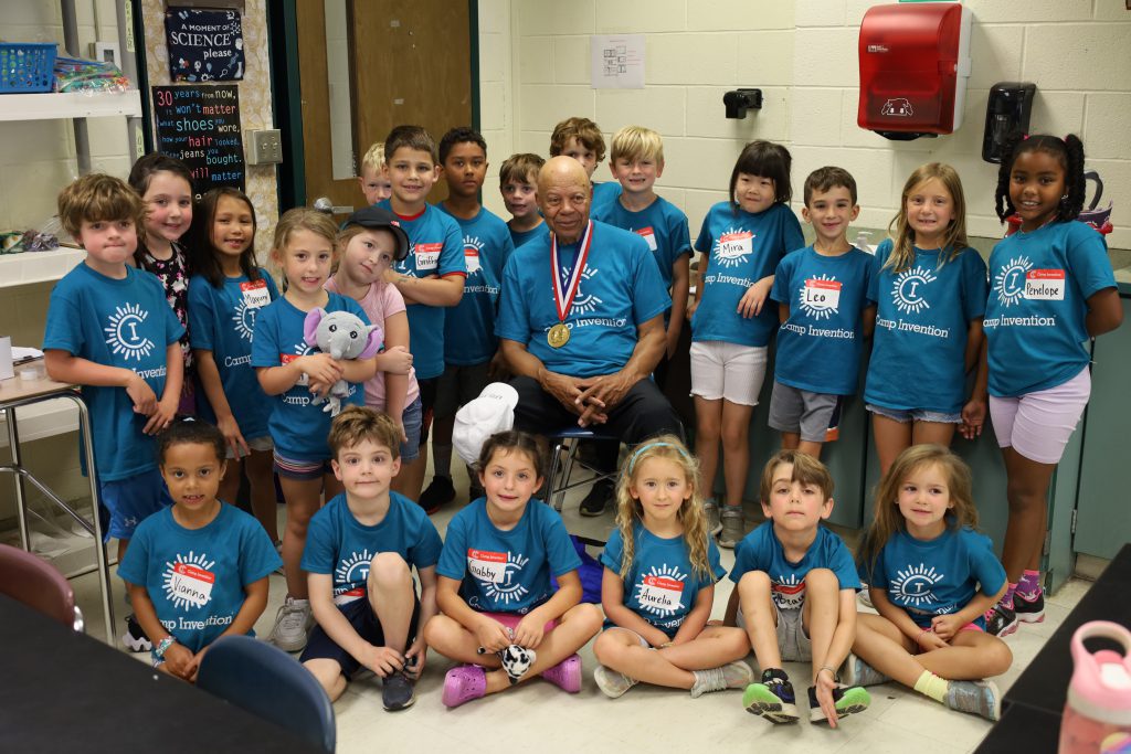 Group photo with elementary students and professional inventor. The inventor is sitting in a chair while the students are standing around him. Some students are sitting on the floor in front.