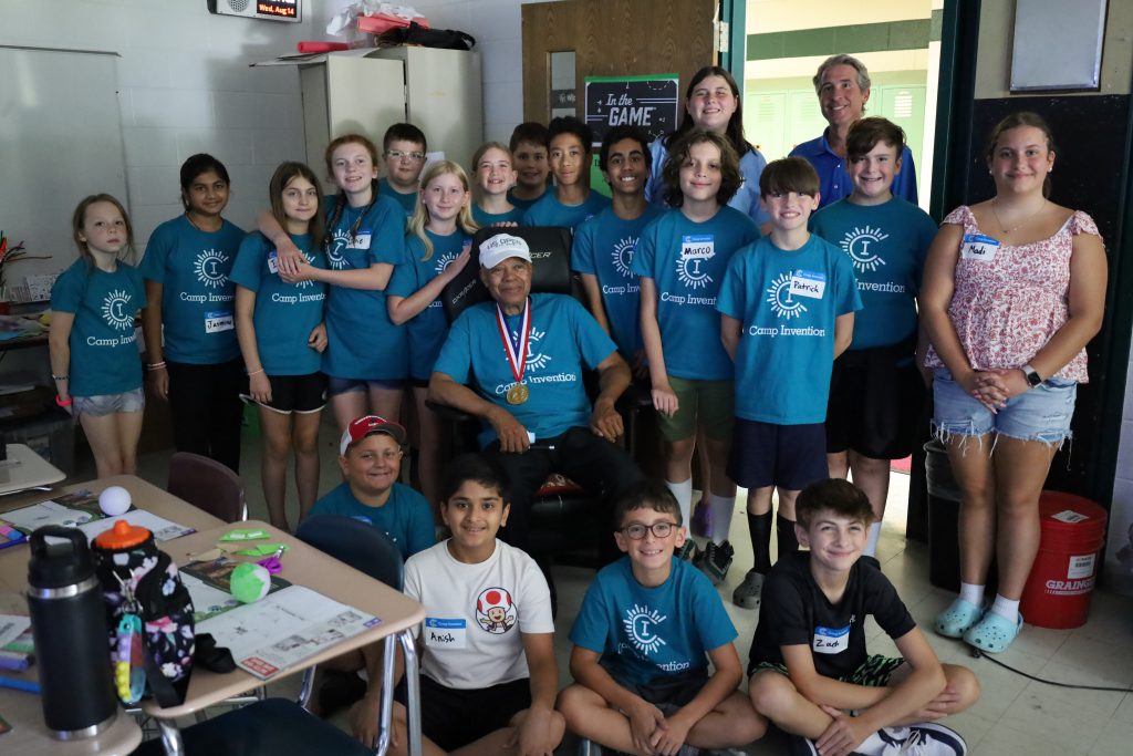 Group photo with elementary students and inventor. The inventor is sitting in a chair while the students are standing around him. Some students are sitting on the floor in front. The school principal is also standing with students in the photo.