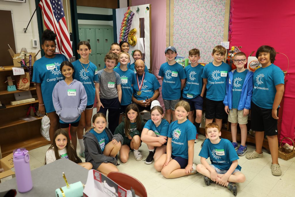 Group photo with inventor and elementary students. The inventor is sitting in a chair while the students are standing behind him. Some students are sitting on the floor in the front.