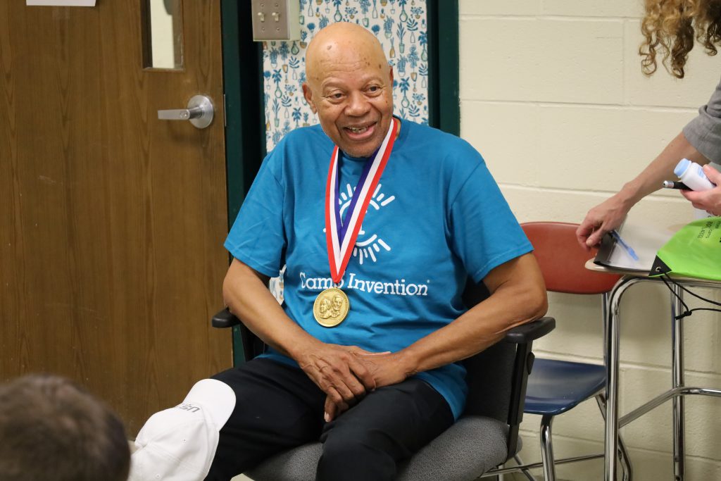 Man sitting in chair smiling with his hands folded in his lap. He has a gold medal around his neck and is smiling.