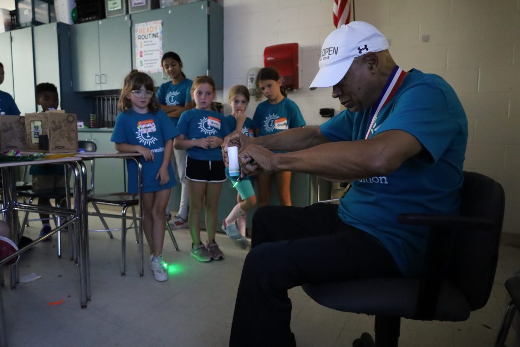 A man is sitting in a chair and sprinkling baby powder while shining a green laser at the same time. Students are standing in the background watching.