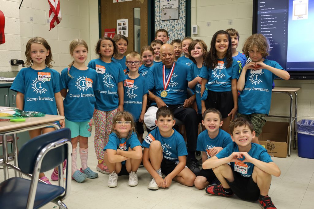 Group photo with inventor and elementary students. The inventor is sitting in a chair while the students are standing around him. Some students are sitting on the floor.