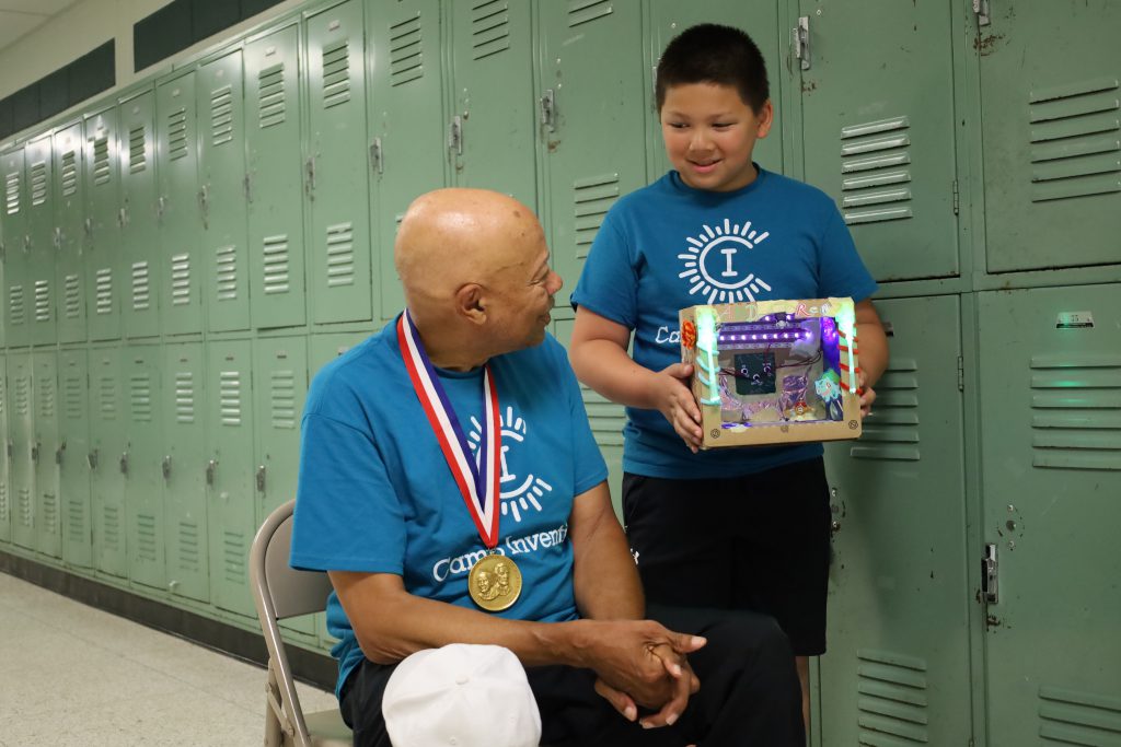 Man sitting in chair with elementary student standing next to him. The student is holding a cardbox electrical glow box and is showing it to the man.