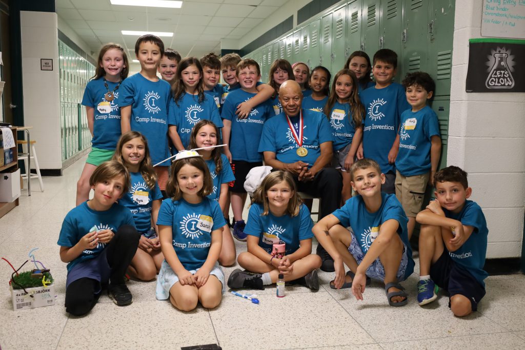 Group photo of inventor and elementary students. The inventor is sitting in a chair while the students are standing around him. Some students are sitting on the floor.