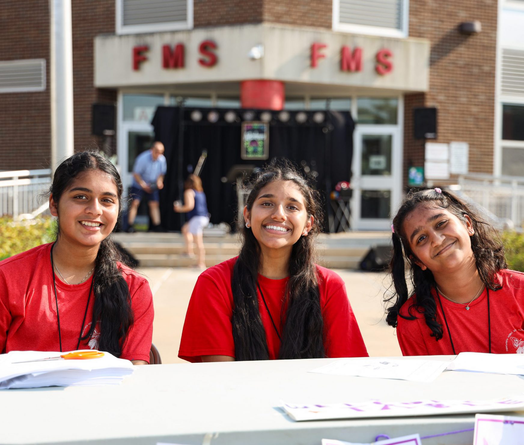 Three students tabling at the Butterfly Station's 25th Anniversary. They are smiling in front of the school.