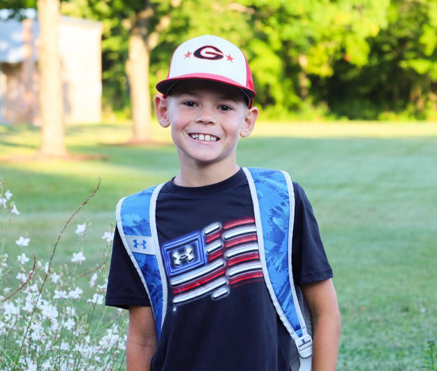 A student wearing a backpack, a t-shirt with a flag graphic on it and a hat with a "G" on it. He is smiling at the camera