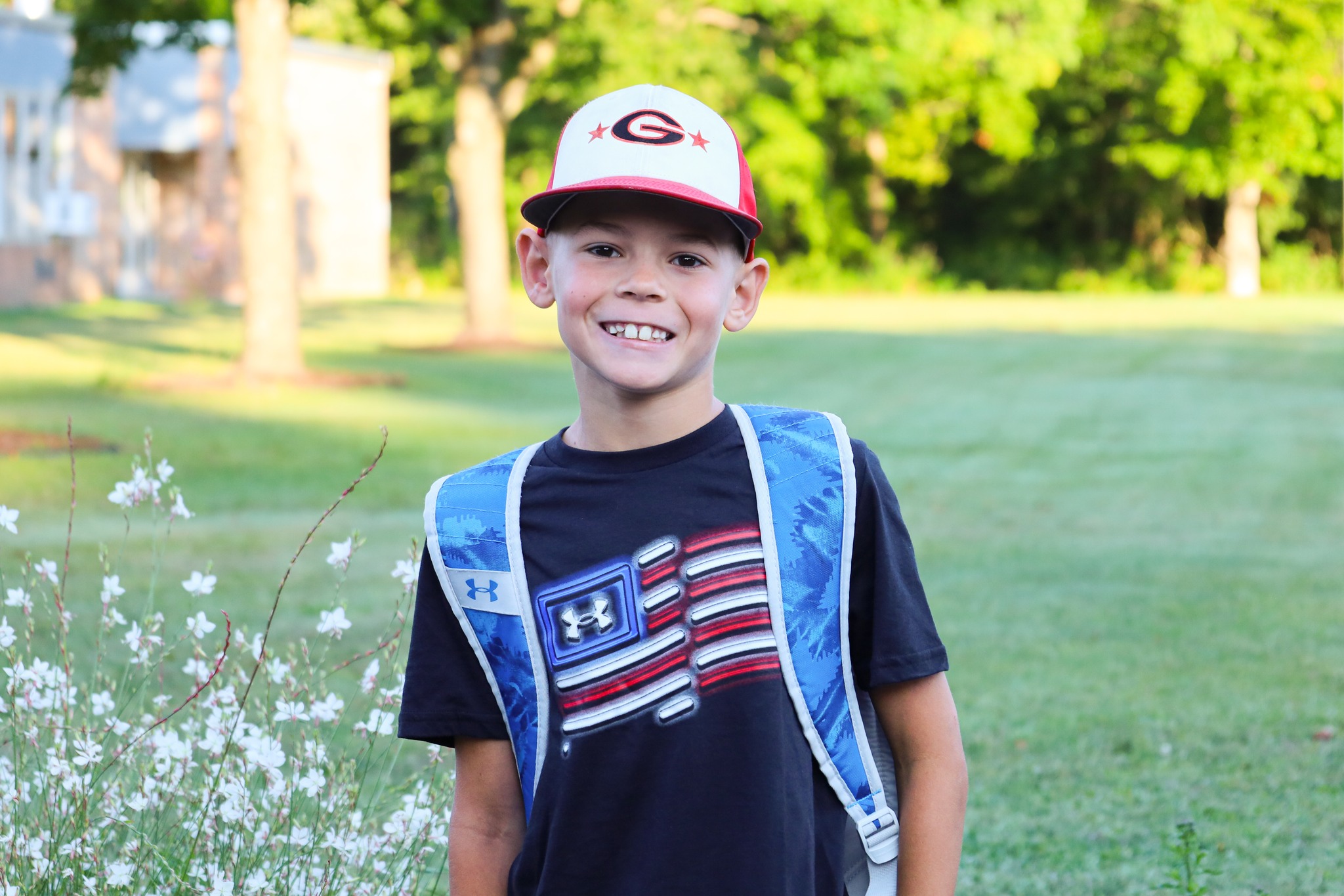 A student wearing a backpack, a t-shirt with a flag graphic on it and a hat with a "G" on it. He is smiling at the camera