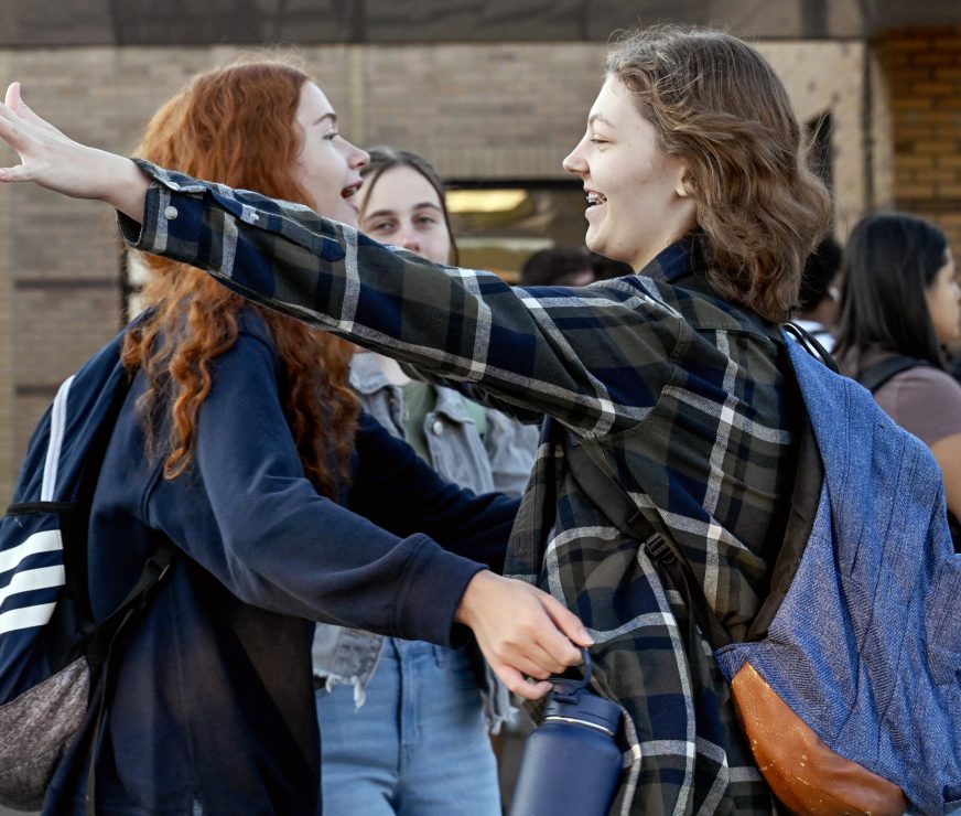 Two students greet each other in front of the high school. They are smiling and getting ready to hug each other