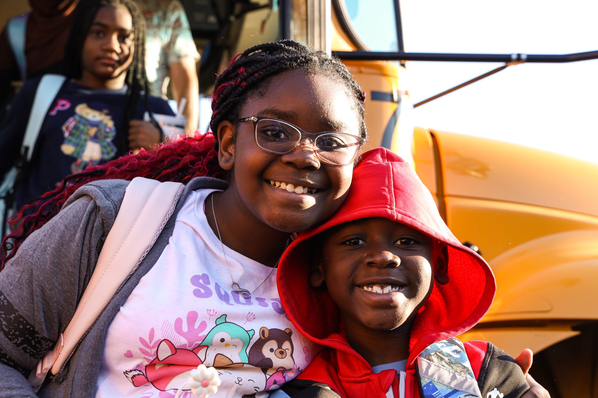 Two students stand in, one has their arm around the other. A school bus is in the background. You can see students disembarking from the bus. The students are smiling