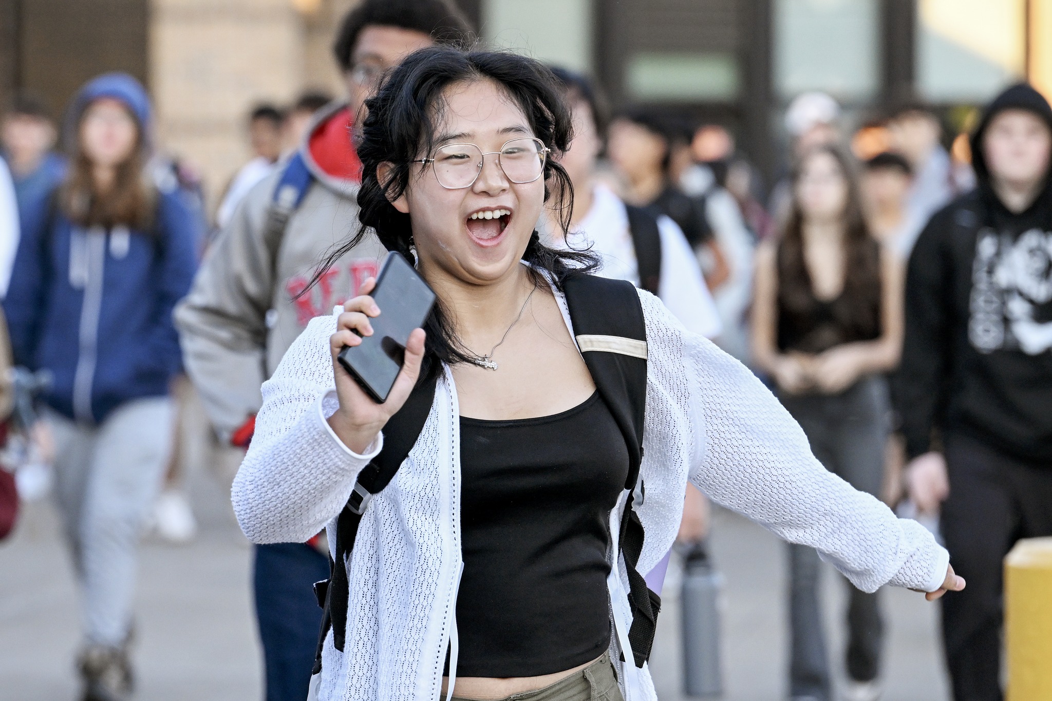 A student is running with their arms open, holding a phone and smiling widely