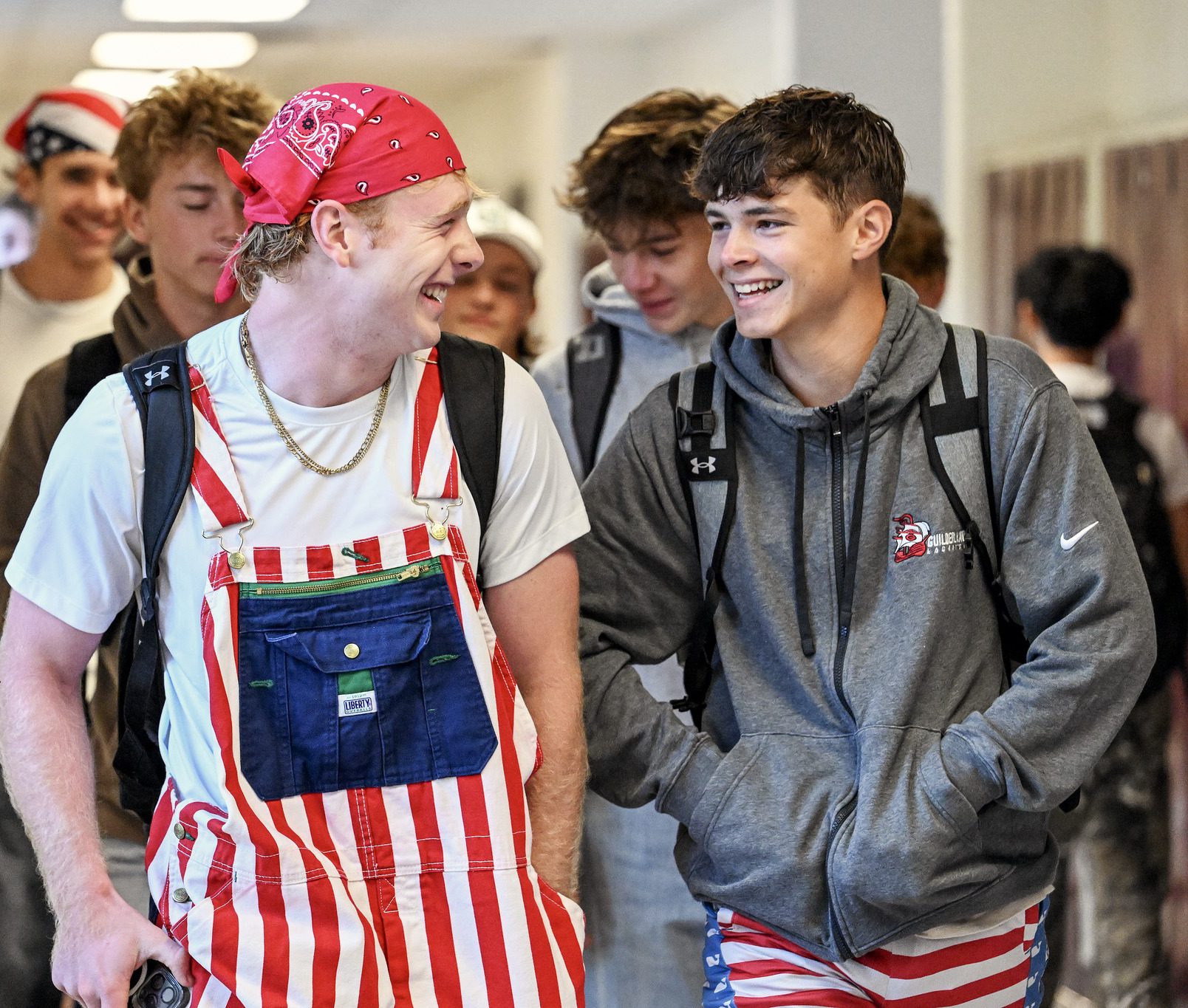 Students are walking down the hallway of the high school. The two students in front of the others are laughing, one is wearing red and white striped overalls and a red bandana on their head, the other has red and white striped shorts and a grey sweatshirt