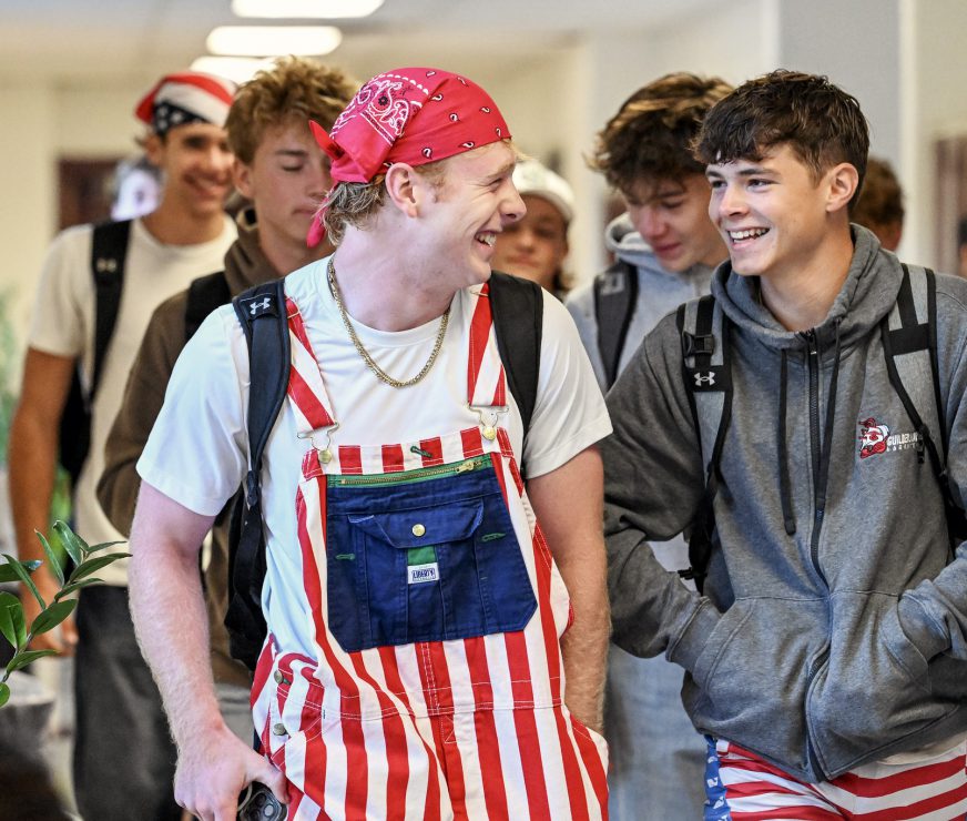 Students are walking down the hallway of the high school. The two students in front of the others are laughing, one is wearing red and white striped overalls and a red bandana on their head, the other has red and white striped shorts and a grey sweatshirt