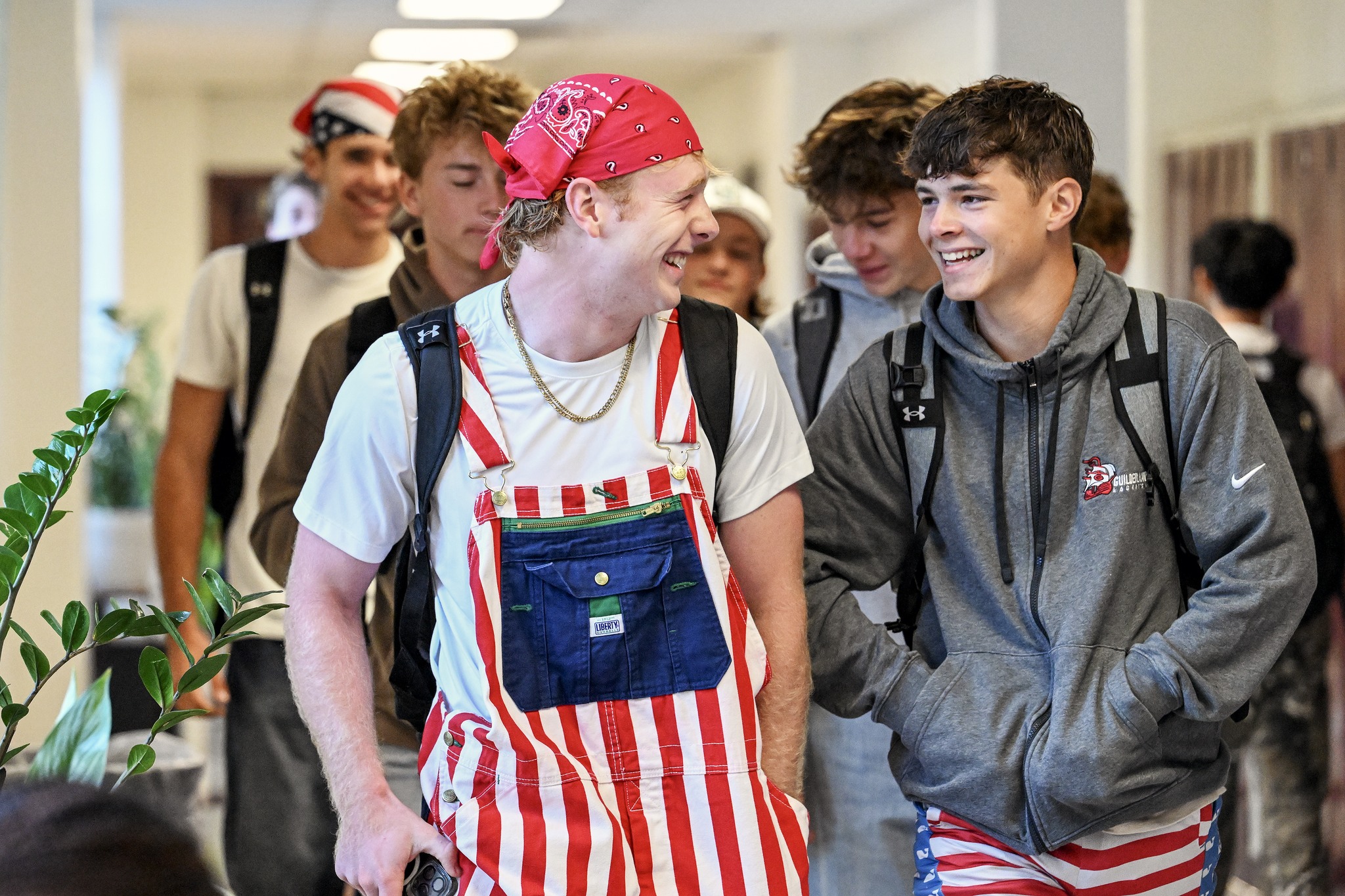 Students are walking down the hallway of the high school. The two students in front of the others are laughing, one is wearing red and white striped overalls and a red bandana on their head, the other has red and white striped shorts and a grey sweatshirt