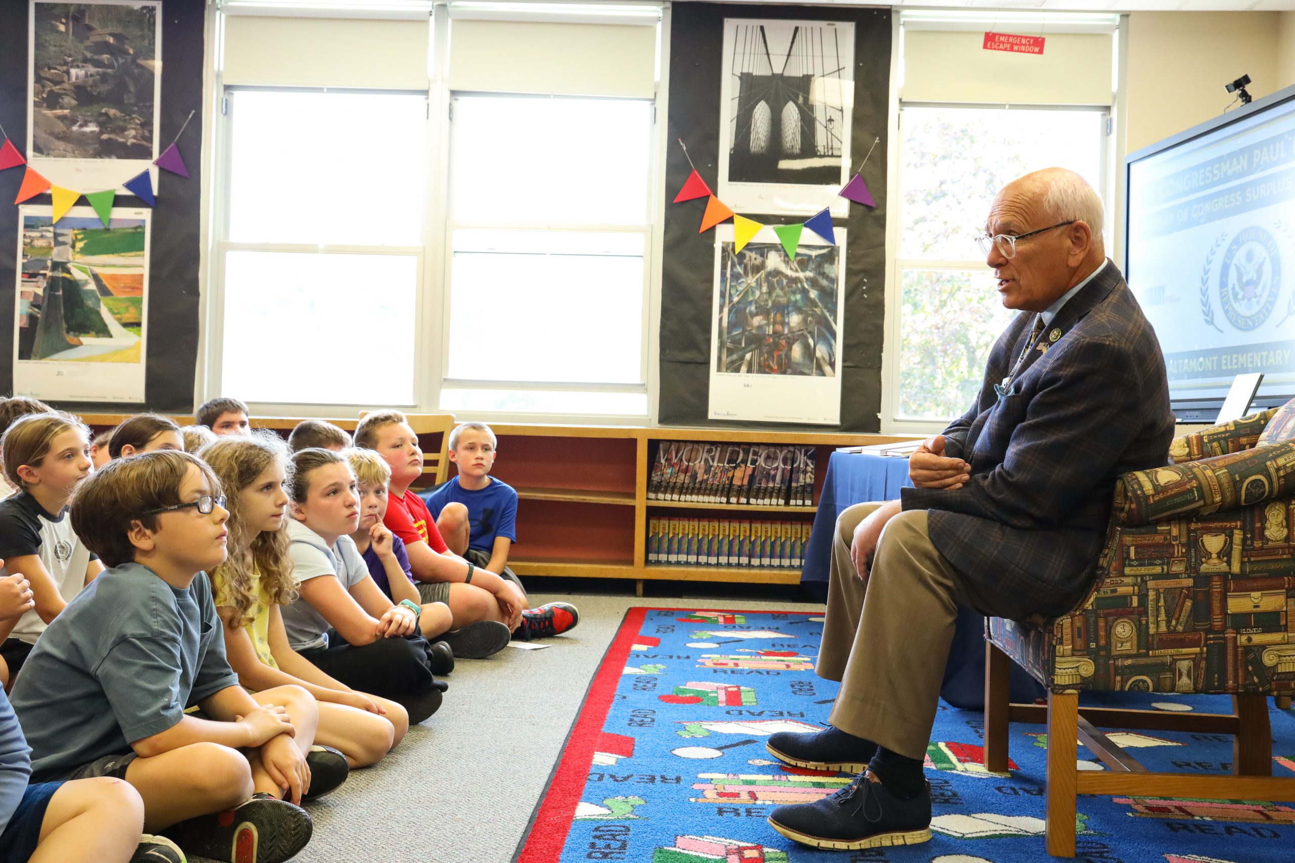 A man sits on a chair. He is facing a group of students who are sitting in rows on the floor. He is speaking to them