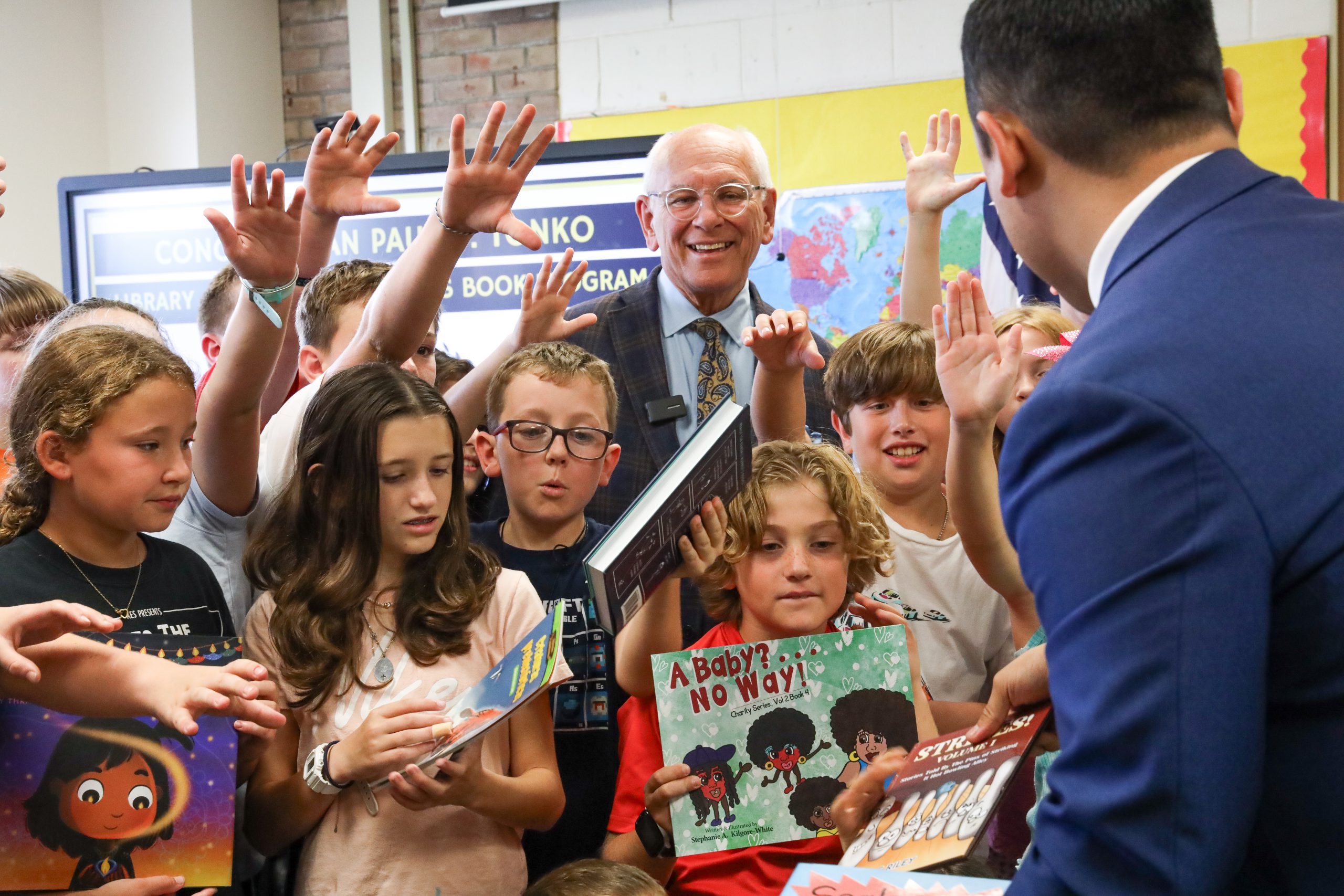 A group of students are eagerly raising their hands and reaching for books, which are being handed out by a person whose back is to the camera. A smiling man stands behind the students.