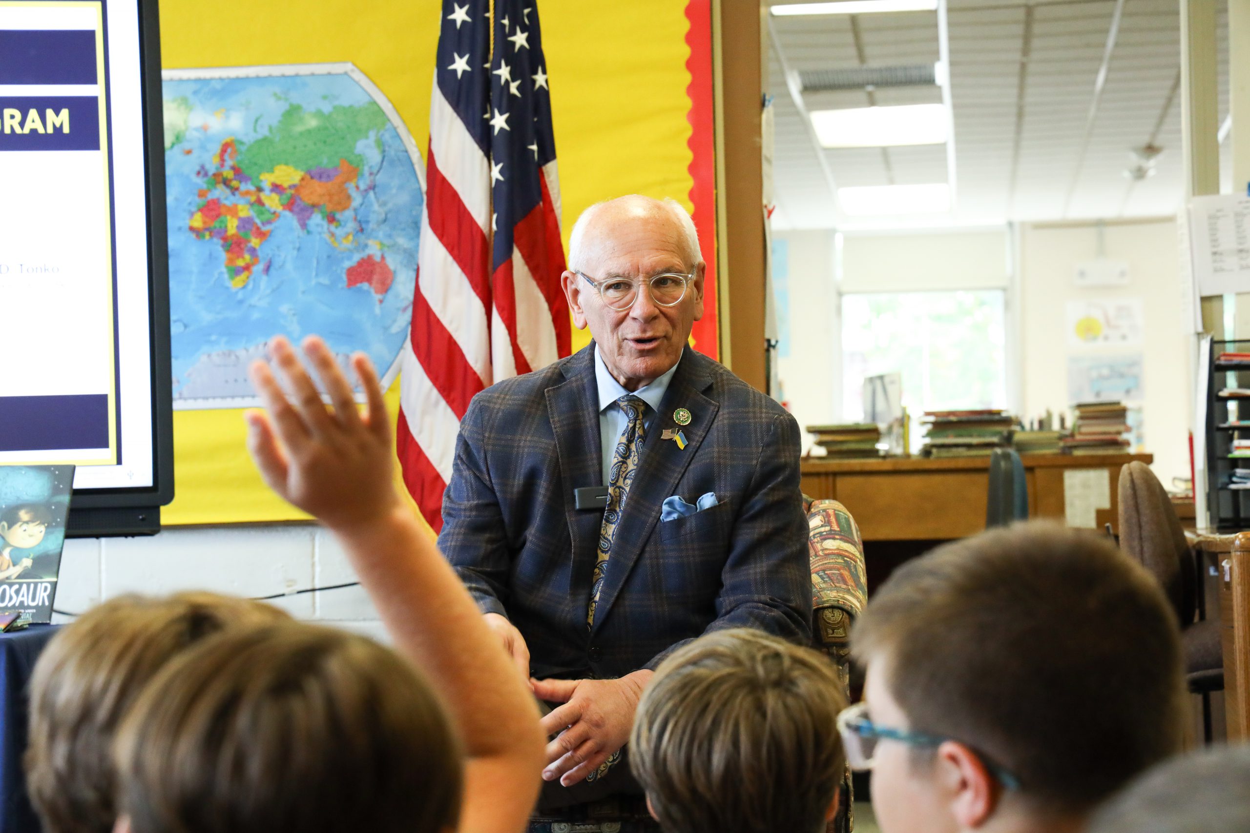 In a school library, a man speaks to a group of students. The photo is taken from behind the students, so you see their heads but not their faces. One student is raising their hand. You can see the man's face who is facing the students. He is wearing a shirt, tie and sport coat