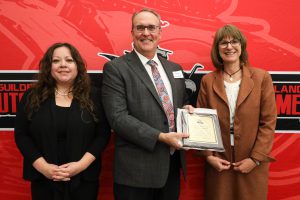 Three people stand in front of a red backdrop. One is holding a plaque. They are smiling at the camera