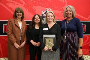 Four people stand in front of a red backdrop. One is holding a plaque. They are smiling at the camera