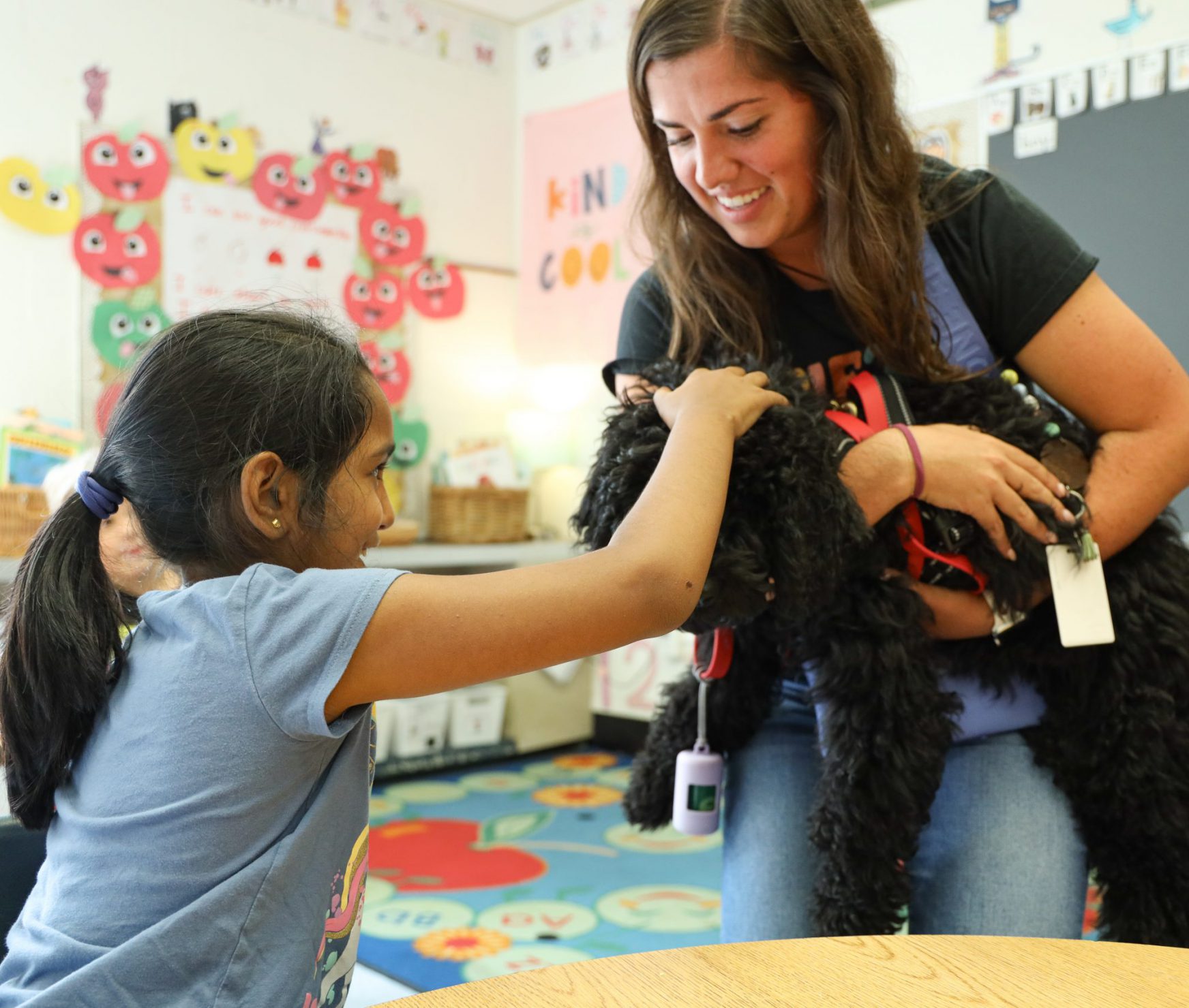 A student is petting a therapy dog. The dog is being held by its handler. The student and handler are smiling.