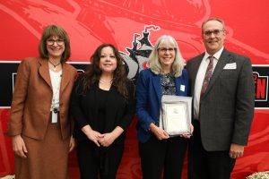 Four people stand in front of a red backdrop. One is holding a plaque. They are smiling at the camera