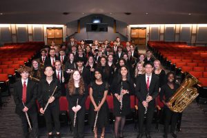 A photo of approximately 60 students, standing in rows in an auditorium. The seats are red, they are wearing black clothing and each is holding a musical instrument