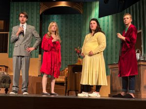 Four students stand on a stage, in costume. There is a set with green old fashioned wallpaper and some furniture. They are wearing costumes. Two are wearing red bath robes, one is in a yellow dress and one is wearing a grey double breasted suit.