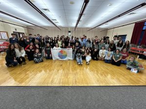 A large group of about 40 students are gathered in the large group instruction room. A few are holding a white banner with a colorful graphic of a wheel divided into 8 sections.