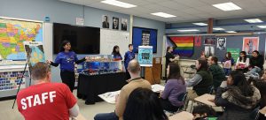 Students stand in the front of a classroom, presenting to a group of people.