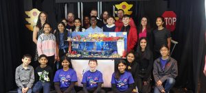 A group of students stand gathered around a model of a city. They are standing in front of a black backdrop.