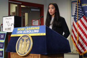 A student stands at a podium, speaking into a microphone. The American flag is behind her