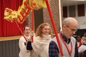 Three teachers participate in a dragon dance. They are holding sticks over their heads. Attached to the sticks is a paper dragon that is yellow and red